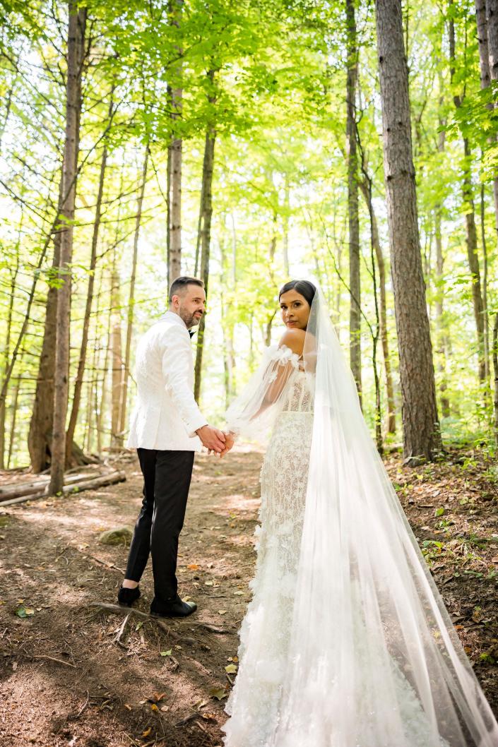 De bruid en bruidegom houden elkaars hand vast en kijken over hun schouders mee in hun trouwkleding terwijl ze in het bos staan.