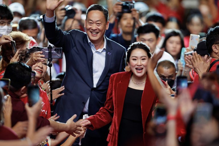 Paetongtarn Shinawatra, dochter van voormalig premier Thaksin Shinawatra, draagt ​​een karmozijnrood fluwelen jasje, een gouden ketting en een zwart wapen.  Ze loopt door de menigte met uitgestrekte handen naar haar toe, sommigen houden hun camera vast.  Ze glimlacht en houdt met haar ene hand haar hand losjes in de menigte die ze voor de andere wil laten vallen.
