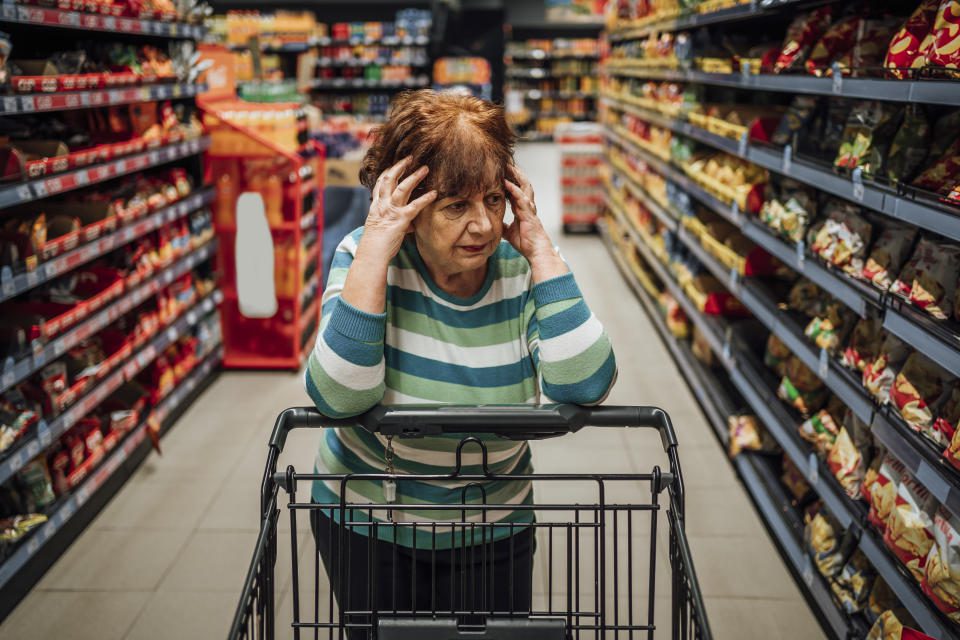 Een oudere vrouw staat in het gangpad met producten in de supermarkt, bezorgd over de stijgende kruideniersprijzen