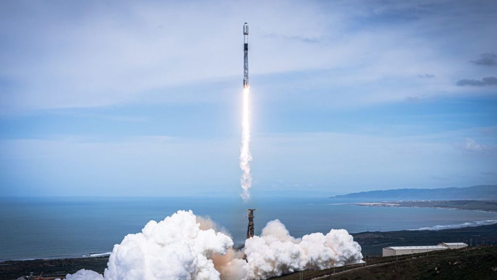 a white and black spacex falcon 9 rocket launches into a blue sky with the ocean in the background.