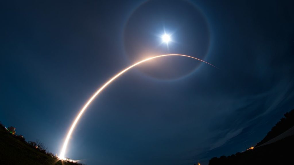a rocket carves a yellow-orange arc into the night sky during a launch.