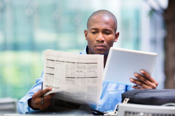Zittende persoon die een financiële krant leest terwijl hij een tablet in zijn linkerhand houdt.